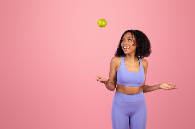 Laughing young african american curly woman in sportswear throws up green apple have fun