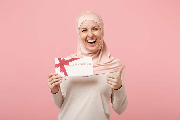 Laughing young arabian muslim woman in hijab light clothes posing isolated on pink background. People religious Islam lifestyle concept. Mock up copy space. Holding gift certificate, showing thumb up.