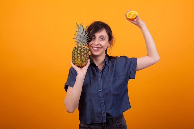 Laughing young beautiful woman with tropical fruits in studio over yellow background. Tasty ananas. Delicious pineapple. Summer fruits.