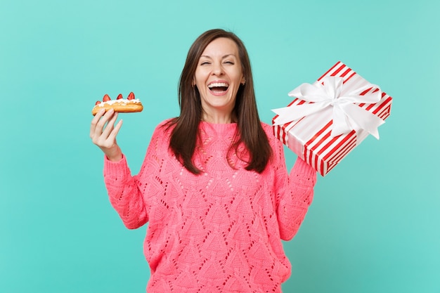 Laughing young girl in knitted pink sweater hold eclair cake red striped present box with gift ribbon isolated on blue background. Valentine's Women's Day birthday holiday concept. Mock up copy space.