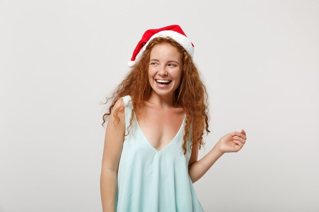 Ridendo giovane rossa santa ragazza in abiti leggeri, cappello di natale isolato su sfondo bianco, ritratto in studio. felice anno nuovo 2020 celebrazione concetto di vacanza. mock up copia spazio. guardando da parte.