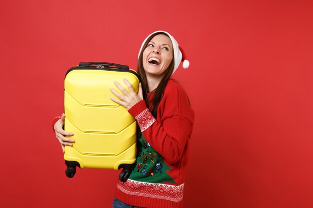 Laughing young Santa girl in knitted sweater, Christmas hat looking up, holding yellow suitcase isolated on red background. Happy New Year 2019 celebration holiday party concept. Mock up copy space.