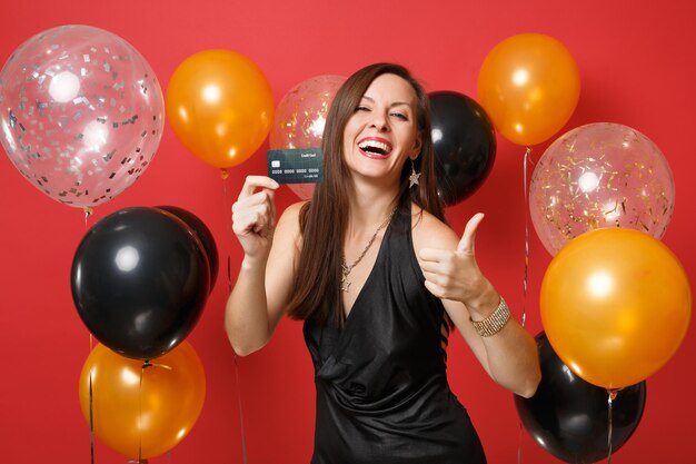 Laughing young woman in black dress celebrating, holding credit card, showing thumb up on red background air balloons. International Women's Day, Happy New Year, birthday mockup holiday party concept.