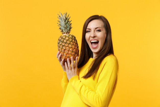 Laughing young woman in casual clothes keeping mouth open, holding fresh ripe pineapple fruit isolated on yellow orange background. People vivid lifestyle, relax vacation concept. Mock up copy space