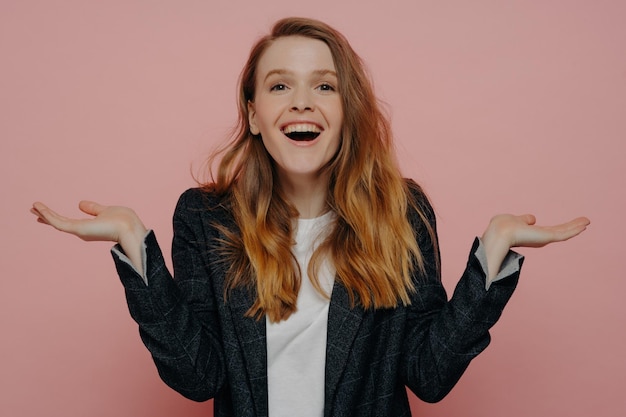 Laughing young woman in dark formal jacket and white top raising hands with open palm gesture looking amused and surprised posing against pink wall