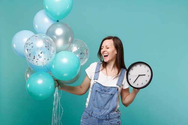 Laughing young woman in denim clothes with closed eyes hold round clock, celebrating with colorful air balloons isolated on blue turquoise background. Birthday holiday party, people emotions concept.