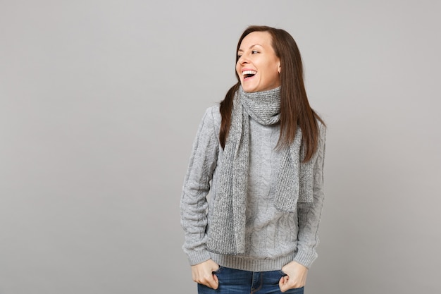 灰色のセーター、灰色の壁の背景のスタジオの肖像画で隔離の脇を探しているスカーフで笑う若い女性。健康的なファッションライフスタイル、人々の誠実な感情、寒い季節のコンセプト。コピースペースをモックアップします。