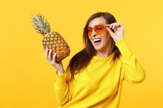 Laughing young woman in heart glasses looking aside hold fresh ripe pineapple fruit isolated on yellow orange background in studio. People vivid lifestyle, relax vacation concept. Mock up copy space.