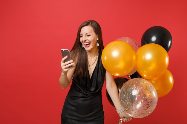 Laughing young woman in little black dress holding air balloons and using mobile phone while celebrating isolated on red background. Women's Day, Happy New Year, birthday mockup holiday party concept.