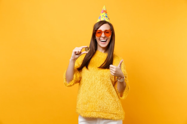 Laughing young woman in orange heart glasses, birthday hat showing thumb up holding bitcoin metal coin of golden color future currency isolated on yellow background. people sincere emotions lifestyle