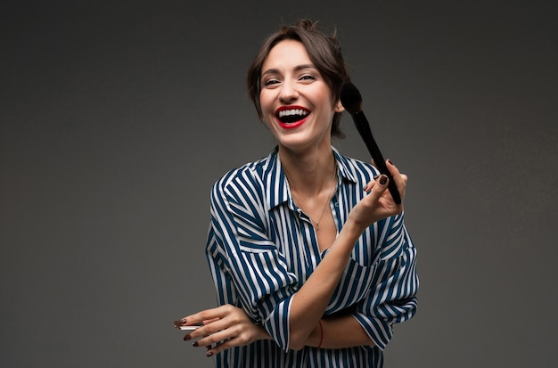 Laughing young woman putting on makeup in striped blouse with makeup brushes in her hands isolated