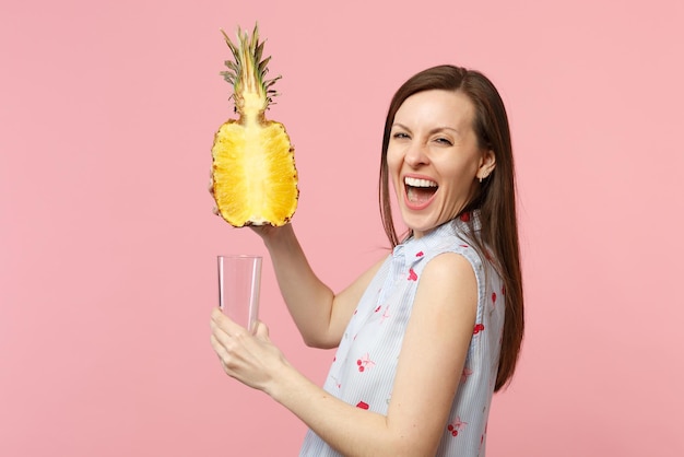 Laughing young woman in summer clothes holding half of fresh ripe pineapple fruit glass cup isolated on pink pastel wall background. People vivid lifestyle, relax vacation concept. Mock up copy space.