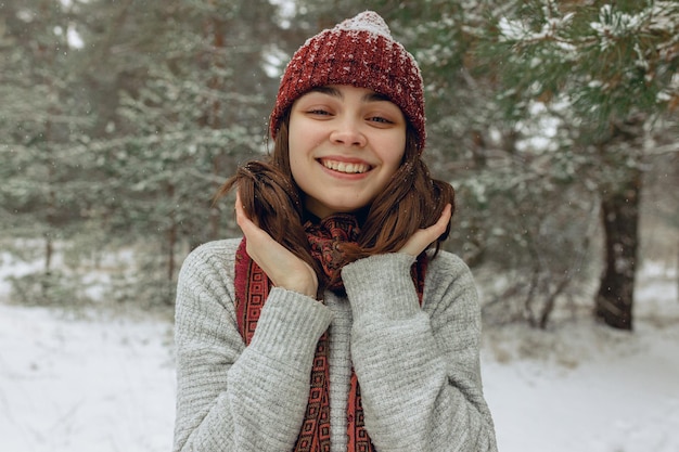 暖かい帽子と雪に覆われた冬の森でカメラを見てスカーフで若い女性を笑ってください。