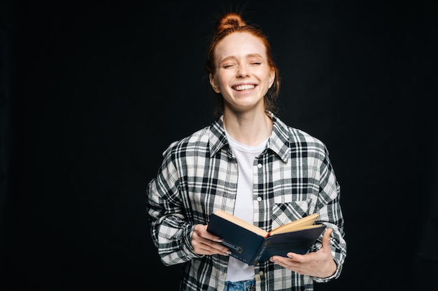 Photo laughing young woman with closed eyes college student holding opened book on black isolated background