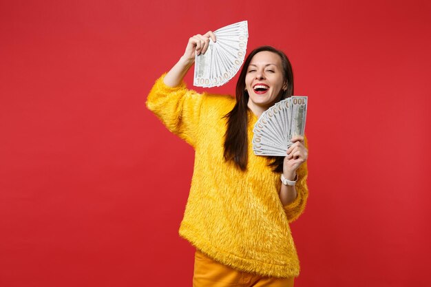 Смеющаяся молодая женщина в желтом меховом свитере, держащая веер денег в долларовых банкнотах, наличные деньги, изолированные на ярко-красном стенном фоне. Люди искренние эмоции, концепция образа жизни. Копируйте пространство для копирования.