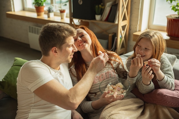 Ridere. la famiglia trascorre del tempo piacevole insieme a casa, sembra felice e allegra. mamma, papà e figlia si divertono, mangiano dolci, guardano la tv. insieme, comfort domestico, amore, concetto di relazioni.