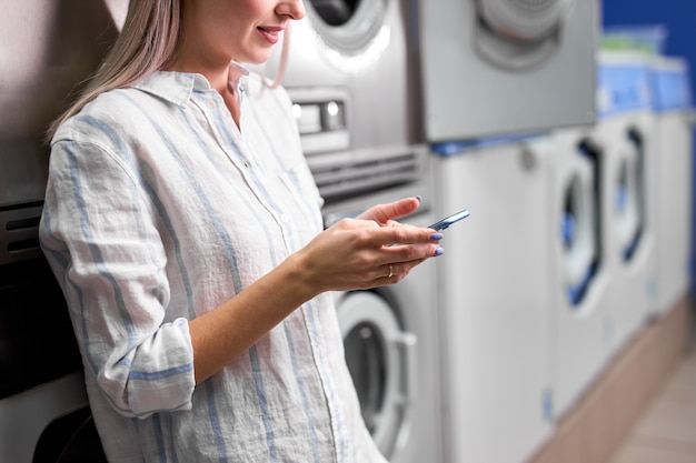 Lavanderia, donna sorridente con telefono in mano, lavare i vestiti tramite app. la giovane femmina caucasica ritagliata è pronta a lavare i vestiti