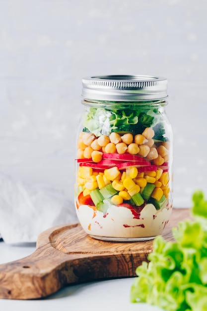 Layering salad in a glass jar Fresh radish cucumber celery red bell pepper corn chickpeas and sauce