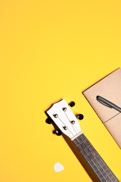 layout with a white ukulele fretboard with nylon strings brown notebook pen and a white pick