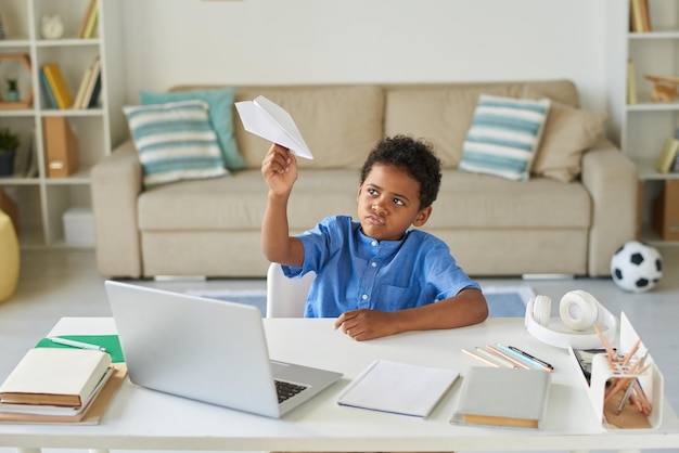 Ленивый черный мальчик с вьющимися волосами сидит за столом и играет с бумажным самолетиком, откладывая на потом