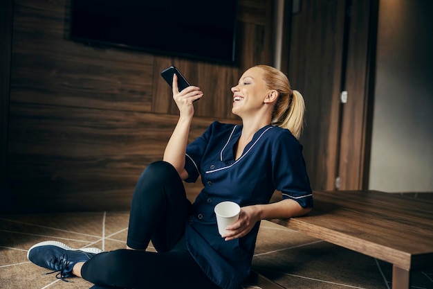 A lazy chambermaid is sitting on the floor and drinking coffee while taking a break and using phone