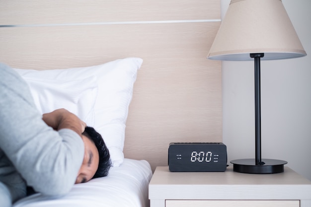 Photo lazy male office worker sleeping on bed in bedroom, cover his ear to block alarm clock alerting sound at 8am in the morning.