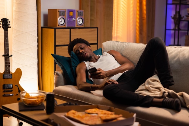 Ленивый мужчина лежит на диване, расслабленный мускулистый афро парень отправляет сообщения, просматривает социальные сети