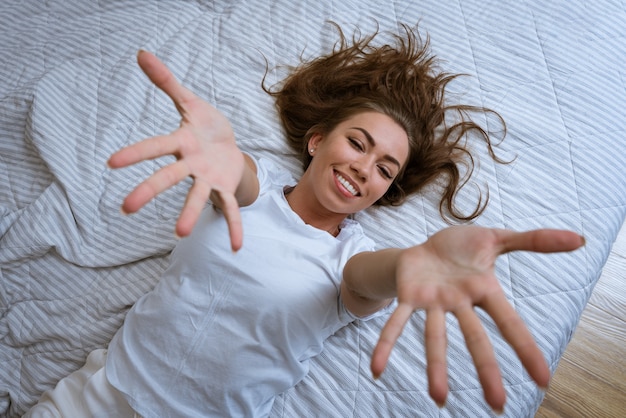 게으른 아침 컨셉의 아름다운 행복한 여성은 침대에 누워서 일어나며 팔짱을 끼고 있습니다.