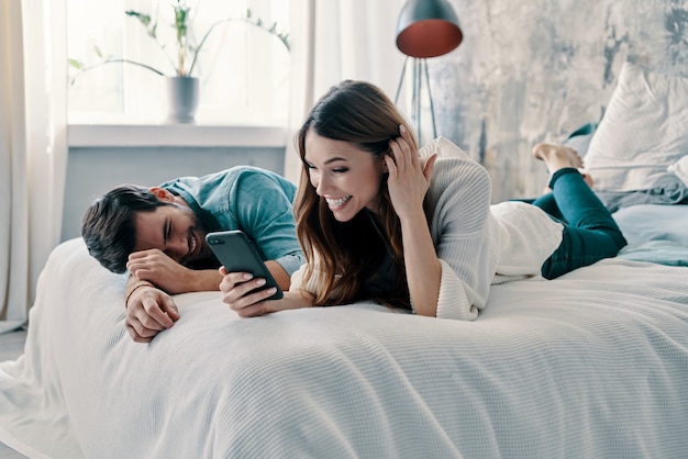게으른 주말. 스마트 폰을 사용하고 집에서 침대에서 시간을 보내는 동안 웃는 아름다운 젊은 부부