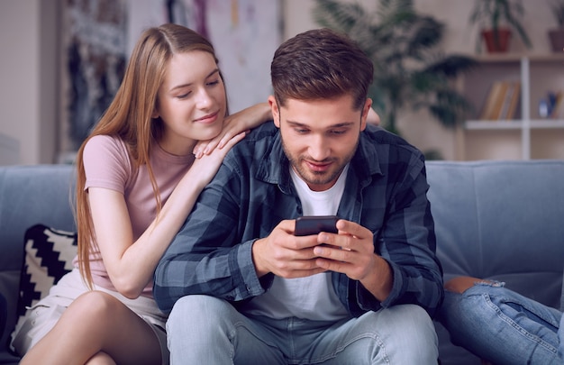 Lazy weekend young beautiful couple in casual wear looking at smartphone while relaxing together on