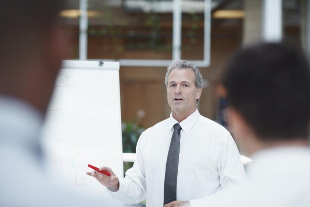 Photo leading the business a senior businessman standing infront of a flipchart with a marker in his hand and talks to his colleagues in the foreground