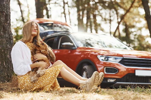 Leaning on the tree. Woman with her dog outdoors in the forest against automobile have good time.