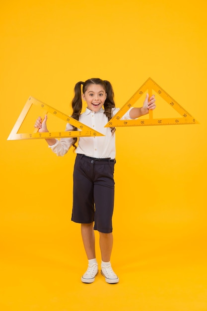 Learning geometry in school School kid holding measuring instruments on yellow background Cute little pupil preparing triangles for school lesson Small girl is back to school