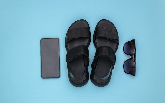 Leather black women's sandals, sunglasses and smartphone on blue background. Top view. Flat lay