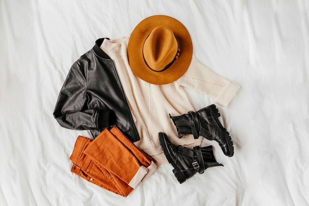 Leather jacket and white warm sweater with brown trousers and hat on white sheet. Women's stylish autumn or winter outfit. Trendy clothes collage. Flat lay, top view.