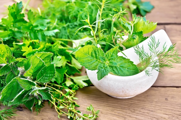 Leaves and sprigs of mint, dill, tarragon, thyme, celery and parsley in a stone mortar with a pestle and on a table against the background of an old wooden board