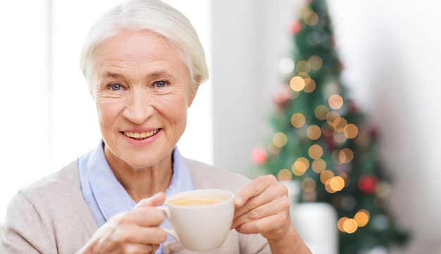 leeftijd, drankje, vakantie, winter en mensen concept - gelukkig lachende senior vrouw met kopje koffie thuis over woonkamer en kerstboom achtergrond