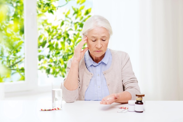 leeftijd, geneeskunde en gezondheidszorgconcept - oudere vrouw met pillen en een glas water thuis op een groene natuurlijke achtergrond