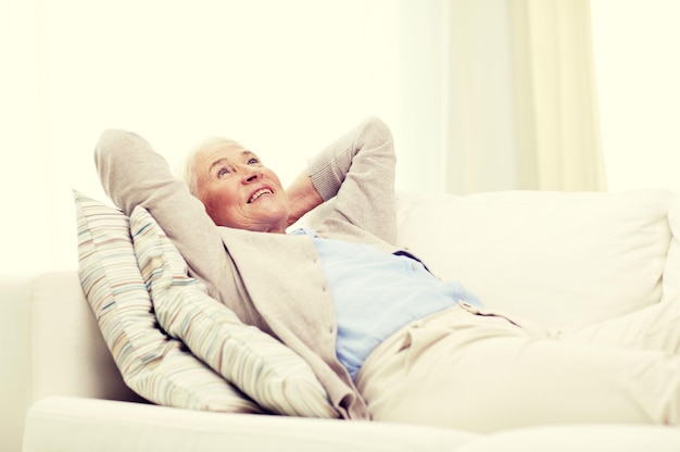 leeftijd, ontspanning en mensenconcept - gelukkig lachende senior vrouw die op de bank rust en thuis droomt