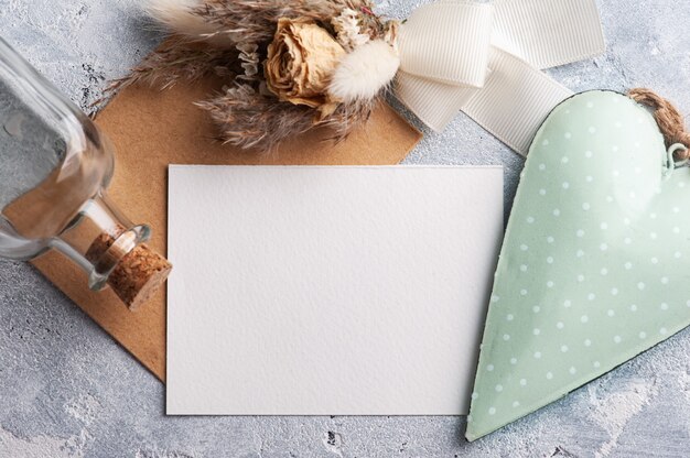 Leeg document en kraftpapier-envelop met groen decoratief hart en droge bloemen. Bruiloft mock up op grijze tafel