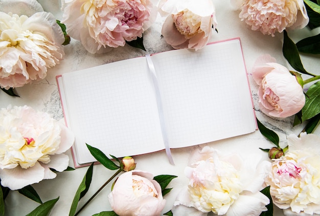 Leeg notitieboekje en roze pioenroos bloemen op witte stenen oppervlak, bovenaanzicht in plat lag stijl