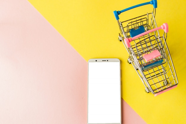 Leeg scherm witte mobiel en winkelwagentjes op pastel roze en geel. Minimale stijl, flatlay