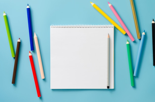 Leeg vierkant notitieboekje en reeks kleurrijke potloden op blauwe achtergrond