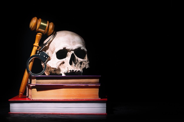 法律、正義、殺人の概念。人間の頭蓋骨と黒い背景に手錠をかけた本に木製の裁判官ガベルハンマー。フリースペース。