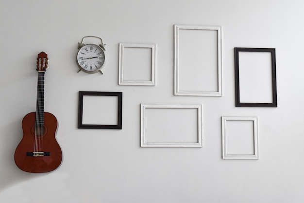 Lege afbeeldingsframes, klok en gitaar op witte cementmuur.