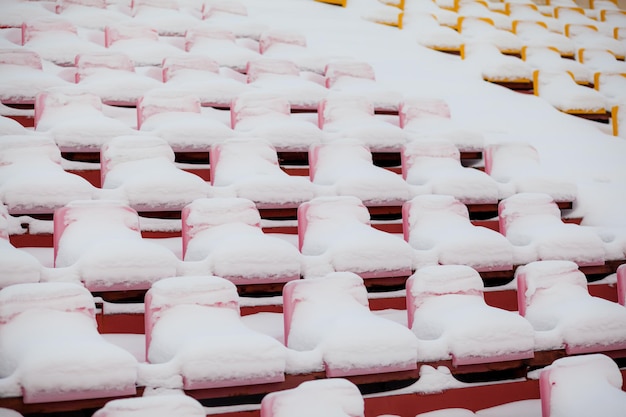 Lege buiten voetbal voetbalstadion stoelen bedekt met sneeuw in de winter, lichte sneeuwval. Plastic stoelen op een rij in het stadion zijn bedekt met sneeuwsneeuw.