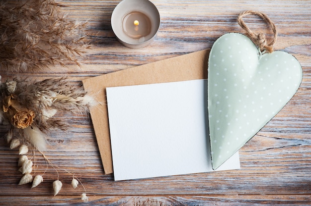 Lege nota en groen decoratief hart, aangestoken aromakaars en droge bloemen op rustieke lijst. Wenskaart voor bruiloft