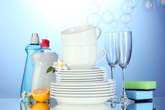 Lege schone borden, glazen en kopjes met afwasmiddelsponzen en citroen op blauwe achtergrond