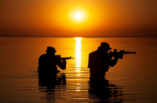 Legermilitairen met het oranje zonsondergangsilhouet van geweren in actie tijdens overval die rivier in het water kruist