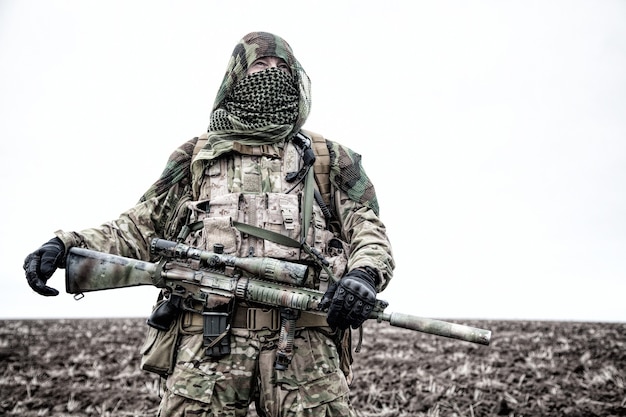 Legerschutter, airsoftspeler in camouflage-uniform en lastdrager, maskerende cape op het hoofd, gewapend dienstgeweer met optisch vizier, verbergend gezicht met shemagh, staand op het veld, in de verte kijkend